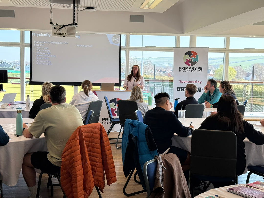 Person giving a presentation at a conference with attendees and a 'Primary PE Conference' sign. The Primary PE Conference is a PE Lead CPD day.