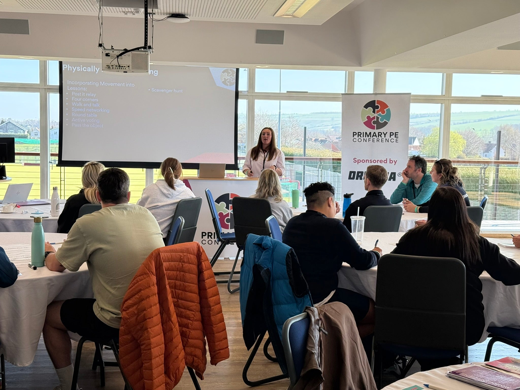 Person giving a presentation at a conference with attendees and a 'Primary PE Conference' sign. The Primary PE Conference is a PE Lead CPD day.