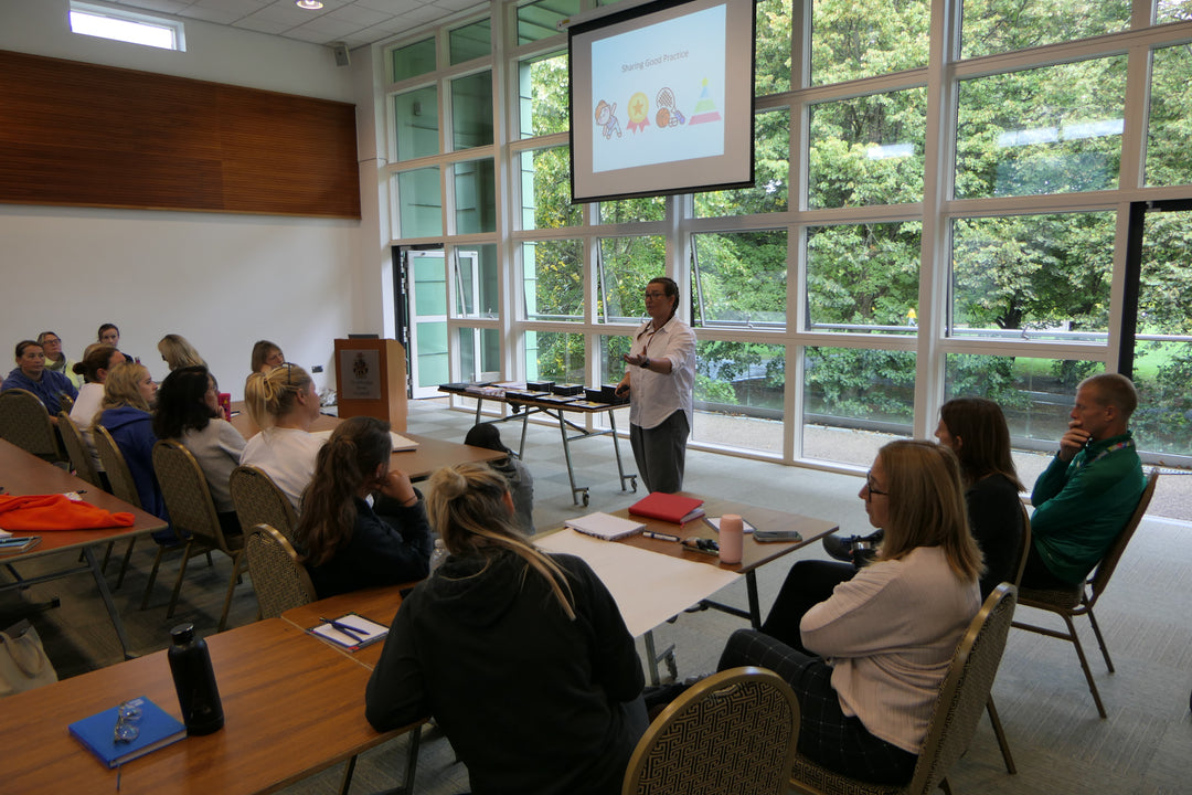 Abbie Morrissey, a woman, giving a presentation in front of people. PE Lead support and consultancy taking place in front of a large window.