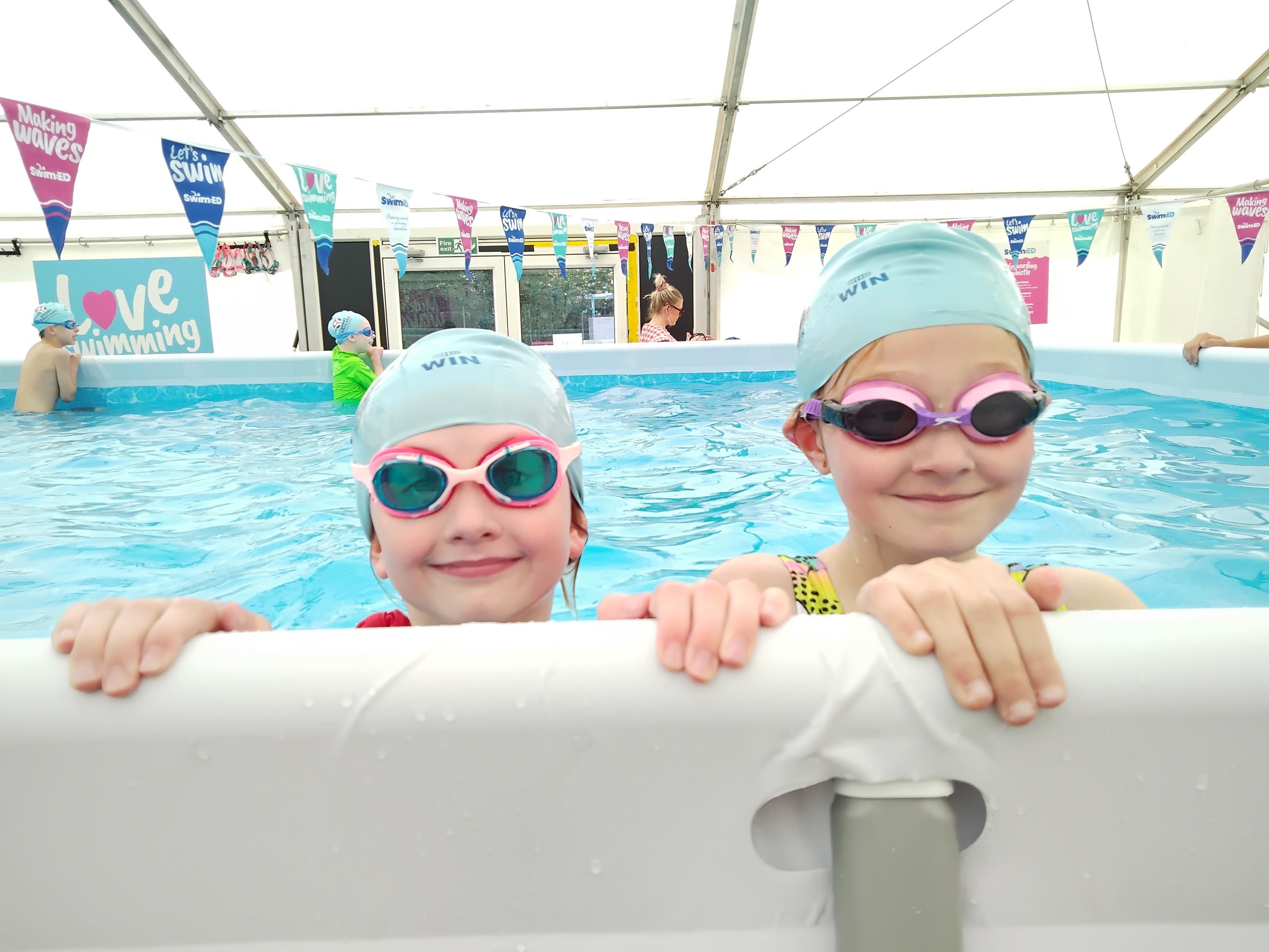 Two children in swim caps and goggles leaning over a pool edge with Swim:ED flags and banners in the background. The portable school swimming pool that gives water confidence for children and ehances primary school swimming lessons. 